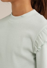 Svijetlozeleni rebrasti pulover s visokim izrezom i volanima na ramenima. Mekana tekstura, uski kroj.