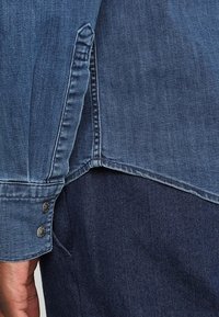 Farkkupaita keskisinisen sävyssä, jossa on napitus hihassa, sivuaukko ja hieman kaareva helma rentoa istuvuutta varten.