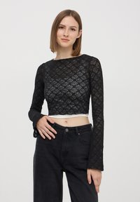 Top a maniche lunghe in pizzo nero corto con motivo floreale, abbinato a pantaloni in denim neri a vita alta. Presenta una maglietta bianca sottostante.