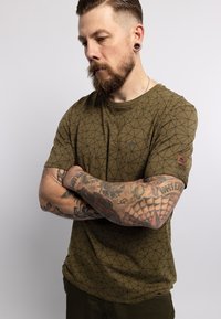 Olijfgroene t-shirt met zwart geometrisch patroon, ronde halslijn, korte mouwen en klein merklogo op de linkermouw. Vloeiende katoenstof.