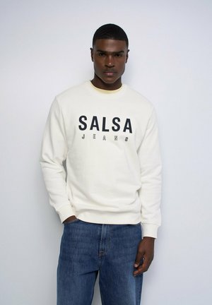 Joven con pelo corto que lleva una sudadera blanca con el texto "SALSA JEANS" y vaqueros azules, de pie frente a un fondo claro y liso.