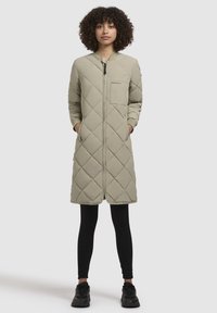 Cappotto lungo trapuntato di colore beige chiaro, caratterizzato da motivi a rombi, con zip frontale completa e tasche laterali. Indossato sopra leggings neri e scarpe.