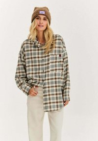 Blondynka w brązowej dzianinowej czapce, oversize'owej szaro-brązowej koszuli w kratę zapinanej na guziki i jasnobeżowych spodniach, stojąca na jednolitym tle.