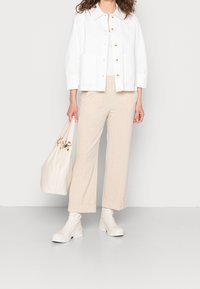 Chaqueta blanca de botones, pantalones beige de pierna ancha con los puños doblados, botas blancas hasta el tobillo y un bolso de mano crema con detalles dorados.