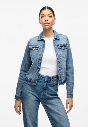 Chaqueta de mezclilla azul claro con cierre de botones, bolsillos en el pecho y mangas largas; combinada con una camisa blanca y jeans azules. Textura suave.