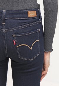 Los jeans de mezclilla azul oscuro presentan un diseño clásico de cinco bolsillos con costuras doradas en contraste y una etiqueta de logo de cuero en la cinturilla.