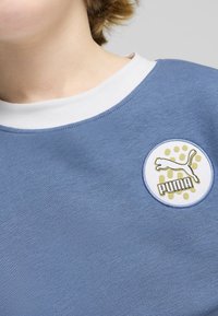 Sudadera azul claro con un cuello blanco, que presenta un logo circular de Puma en blanco y dorado con un fondo de puntos. Textura suave.