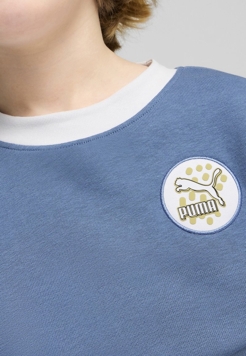 Sudadera azul claro con un cuello blanco, que presenta un logo circular de Puma en blanco y dorado con un fondo de puntos. Textura suave.