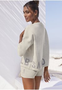Maglione in cotone grigio chiaro con vestibilità ampia e logo "ELB SAND". Abbinato a pantaloni corti coordinati, con una texture morbida e un design casual, sullo sfondo di una spiaggia.