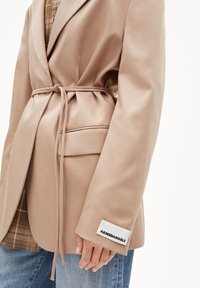 Blazer beige con cintura a cravatta, maniche lunghe, tessuto testurizzato, tasca frontale e etichetta bianca con la scritta "ARMEDANGELS" sul polsino.