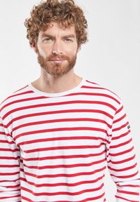 T-shirt a maniche lunghe a righe rosse e bianche con scollatura a girocollo. Materiale in cotone, texture liscia, con un piccolo logo sulla manica sinistra.