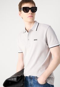 Camisa polo em cinza claro com detalhes em preto e logotipo, com mangas curtas e colarinho clássico, combinada com óculos de sol pretos em formato quadrado e jeans de denim.