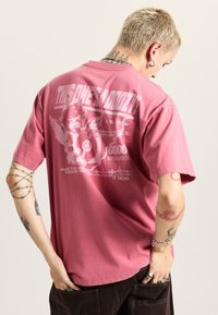 Lyserød t-shirt i bomuld med grafisk tryk på ryggen, herunder tekst og en illustration med englevinger og et design af tornebelte.
