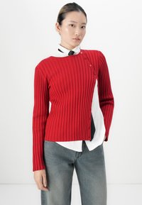Jonge vrouw met samengebonden haar, gekleed in een rode ribgebreide trui met knopen over een wit shirt en zwarte stropdas, gecombineerd met blauwe jeans.