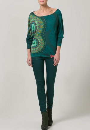 Frau trägt ein tealfarbenes, schulterfreies Langarmoberteil mit farbigem Mandala-Design sowie passende schmale tealfarbene Hosen und Stiefel.