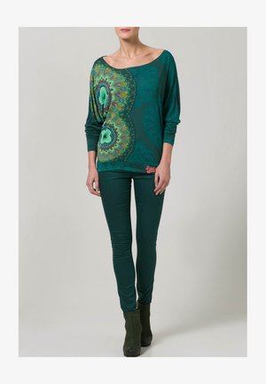 Kvinde iført teal skulderløs langærmet top med farverigt mandala-design og matchende teal smalle bukser og støvler.
