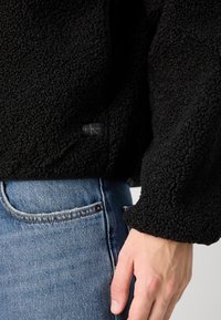 Giacca in pile nera con superficie morbida e testurizzata; presenta una toppa con logo visibile ed è indossata con jeans di denim blu.