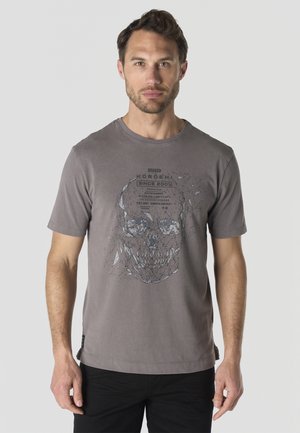 Hombre de pie con camiseta gris con un gráfico geométrico de calavera y pantalones negros sobre un fondo liso.