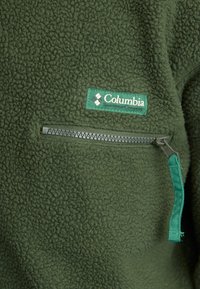 Green fleece jacket section with Columbia Sportswear logo patch and horizontal zipper pocket with fabric pull tab.