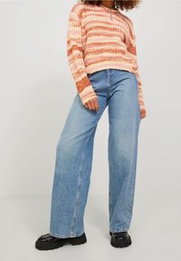 Pletený svetr s růžovými a oranžovými pruhy, doplněný světle modrými kalhotami širokého střihu a černými robustními botami. Texturovaný a ležérní vzhled.