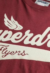 Czerwona bawełniana koszulka z dużym białym logo "Superdry" z projektem skrzydeł powyżej i napisem "Flyers" poniżej w kursywie. Miękki materiał, okrągły dekolt.