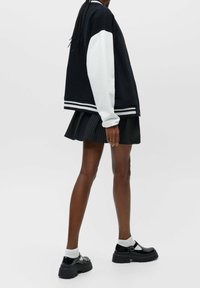 Black and white varsity jacket with drop shoulders, striped ribbed cuffs, and a relaxed fit, paired with a pleated black skirt and chunky shoes.