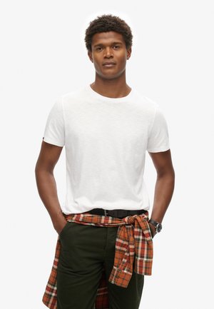 Hvid t-shirt med korte ærmer parret med olivengrønne bukser. En orange og ternet skjorte er bundet om taljen. Sort bælte og ur fremhæver outfittet.