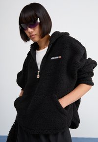 Haut à capuche en polaire noire avec fermeture éclair à l'avant, grandes poches et détail de logo. Le tissu a une apparence texturée et duveteuse.