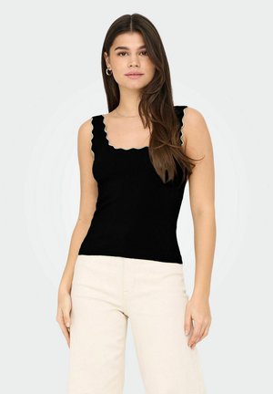 Joven mujer con cabello largo y castaño, vistiendo una blusa negra sin mangas con borde ondulado y pantalones beige claros, de pie frente a un fondo liso.