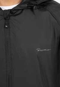 Zwarte lichtgewicht jas met een rits aan de voorkant, verstelbare kap en een minimalistisch wit logo op de borst. Soepele textuur, eenvoudig ontwerp.