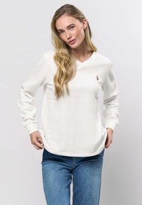 Vit långärmad V-ringad sweatshirt med ribbade muddar och fåll, med en liten broderad detalj på bröstet. Bärs med blå jeans.