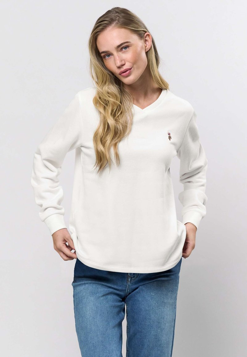 Vit långärmad V-ringad sweatshirt med ribbade muddar och fåll, med en liten broderad detalj på bröstet. Bärs med blå jeans.