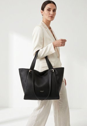 Donna in abito chiaro che porta una grande borsa a mano in pelle nera con tracolle, in piedi contro uno sfondo bianco uniforme.