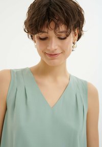 Blusa senza maniche verde chiaro con scollatura a V e pieghe sottili sulle spalle, realizzata in tessuto morbido. Design semplice e neutro.