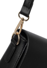 Detalle de un bolso de cuero negro con superficie texturizada, clip de metal dorado y un punto de sujeción para la correa. Bordes suaves con costuras precisas.