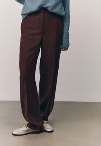 Pantaloni a gamba larga in tessuto bordeaux, con cuciture verticali e texture liscia, abbinati a mocassini di colore chiaro.