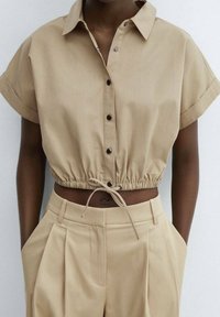 Camicetta cropped beige con bottone frontale, maniche corte e vita elasticizzata. Abbinata a pantaloni beige a vita alta.