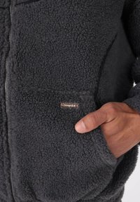 Mano parzialmente nella tasca anteriore di una giacca in fleece grigio scuro, con una texture evidente e una cerniera visibile lungo il lato.