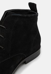 Botine negre din suede, cu o textură netedă, vârf ascuțit, design cu șireturi și un tab de prindere în spate pentru a facilita încălțarea.