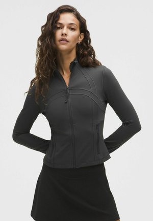 Kobieta z długimi kręconymi włosami, ubrana w dopasowaną czarną kurtkę na suwak i czarną spódnicę, stojąca z rękami za plecami.