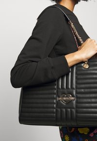 Modell klädd i en svart sweatshirt, som håller en svart vadderad handväska med en gyllene hjärtlogo. Färgglada mönstrade byxor är delvis synliga.