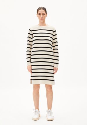 Strickkleid mit horizontalen schwarzen Streifen auf einem cremefarbenen Hintergrund, Rundhalsausschnitt, langen Ärmeln und seitlichen Schlitzen. Kombiniert mit weißen Sneakers.