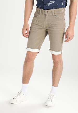 Mand iført beige opkørte shorts, hvide sneakers, hvide sokker og en mørkeblå skjorte med blomsterprint, der står mod en hvid baggrund.
