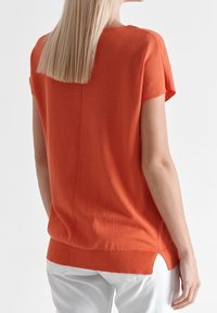 Suéter naranja de manga corta con cuello redondo, aberturas laterales y una costura cosida que detalla la parte posterior. Tejido suave y ligero.