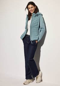 Giacca imbottita azzurro chiaro con cappuccio, abbinata a jeans di denim scuri e sneakers beige. La giacca presenta una cerniera e pannelli testurizzati.