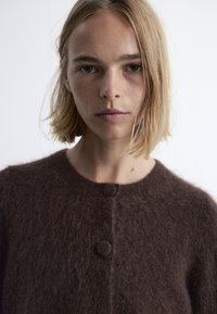 Braune, flauschige Strickjacke mit Rundhalsausschnitt, zwei Knöpfen und weicher Textur; Modell steht frontal, minimaler Hintergrund.