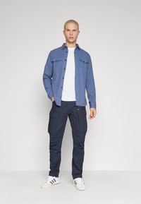 Cămășă albastră cu nasturi și buzunare pe piept, tricou alb cu guler rotund, pantaloni cargo negri cu fermoar și buzunare, și adidași albi cu dungi.