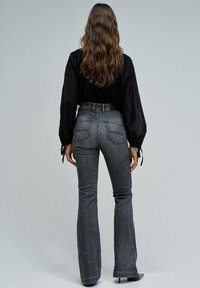 Kobieta z długimi falującymi włosami, ubrana w czarną bluzkę i szare dzwony z wysokim stanem, stojąca tyłem na jednolitym tle.