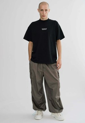 Black short-sleeve t-shirt with white logo, paired with loose-fitting, light brown cargo pants and white sneakers. Plain background.