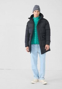 Hombre con un gorro gris, abrigo de plumas negro, chaqueta turquesa, jeans azul claro y zapatillas blancas, de pie frente a un fondo claro y liso.